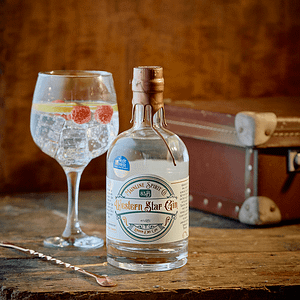 Bottle shot of a 70cl, Western Star Gin. Served in a balloon glass with tonic, ice and a slice of lemon with a raspberry. Sat on an old table with a vintage suitcase in distance.